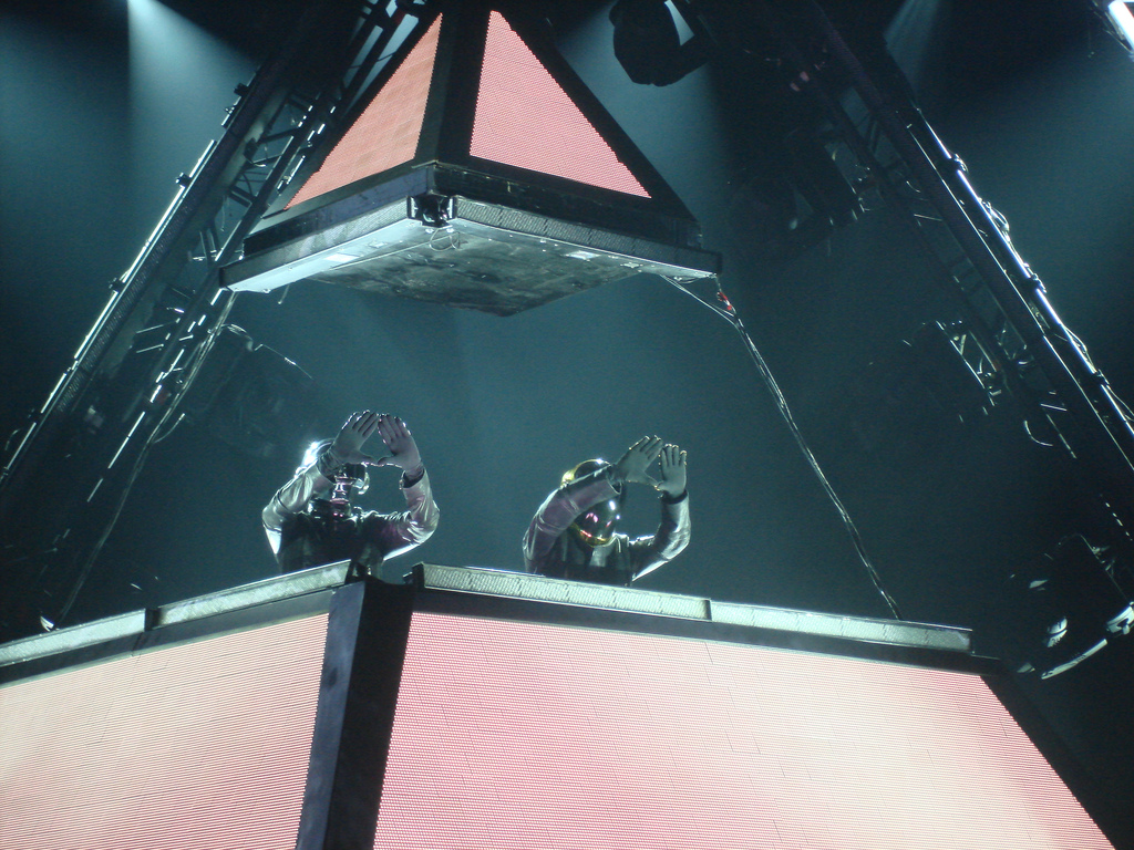Daft Punk performing live in their illuminated pyramid, raising their hands in the air to form a pyramid shape with their hands.