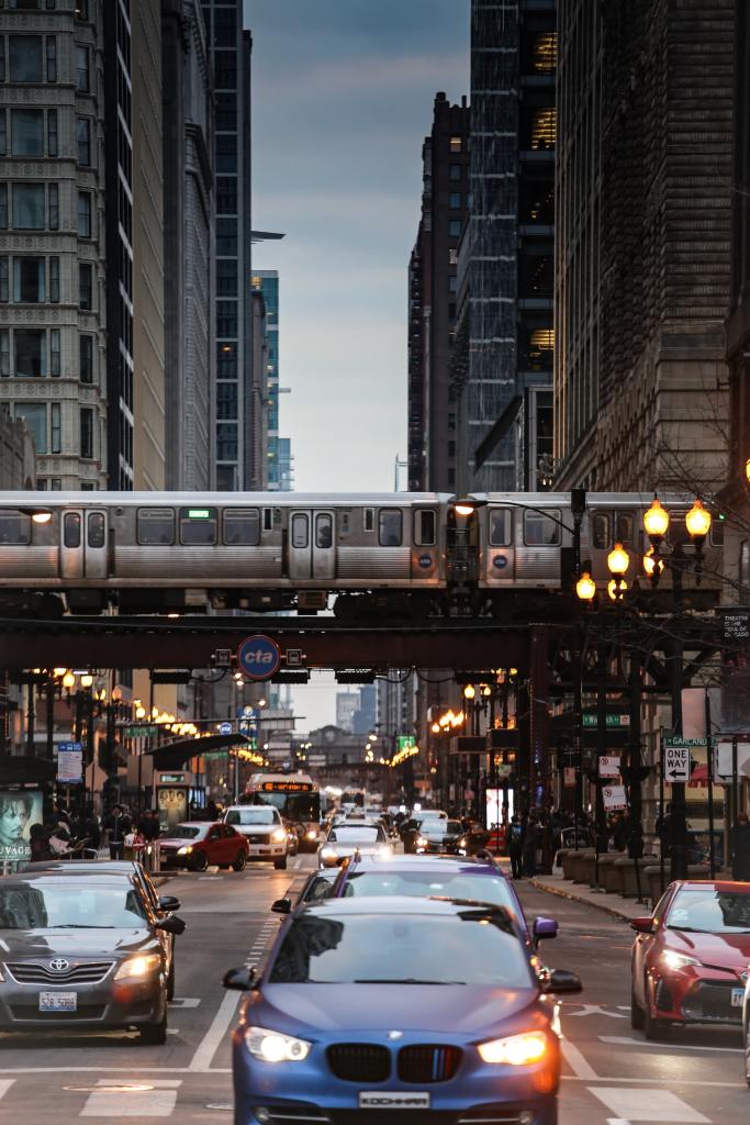 Cars driving through a busy city street with a passenger train passing over.