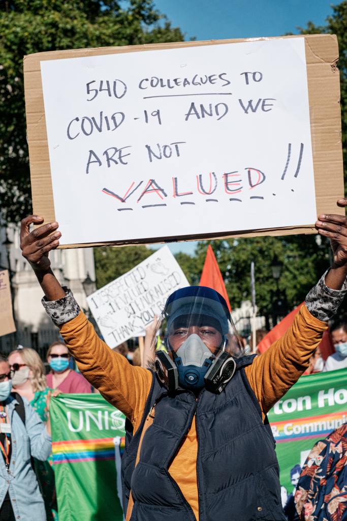 Woman in orange and black jacket holding white printer paper that states "540 collagues to covid-19 and we are not valued"